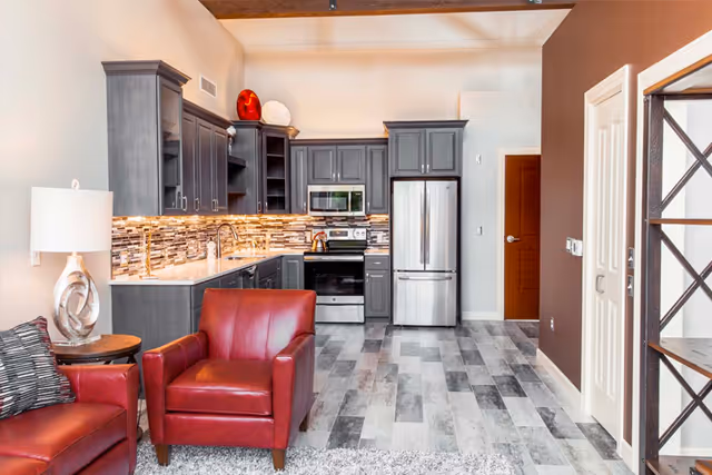 Open-plan living area with red leather chairs in front of a modern kitchen featuring gray cabinets and stainless steel appliances.