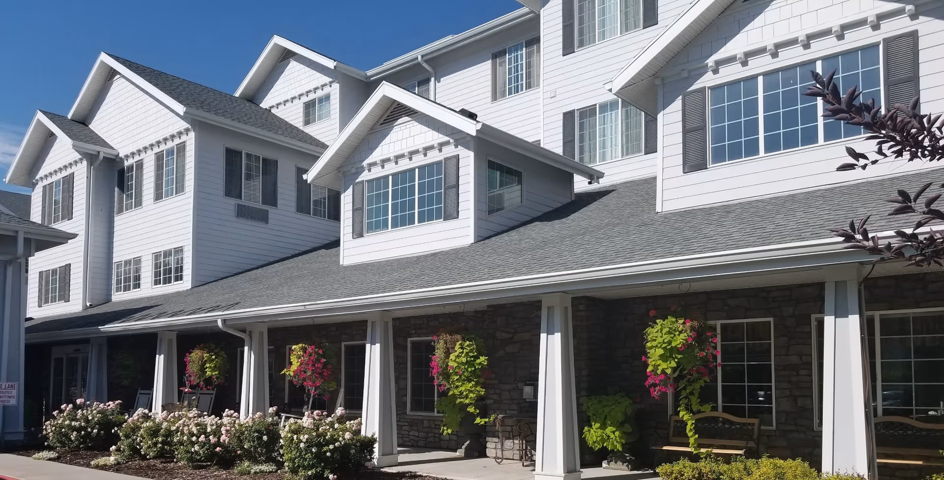 Exterior view of a multi-story senior living facility building with white siding and stone accents. The building has several large windows and a covered porch area with hanging flower baskets and benches. The sky is clear and blue.