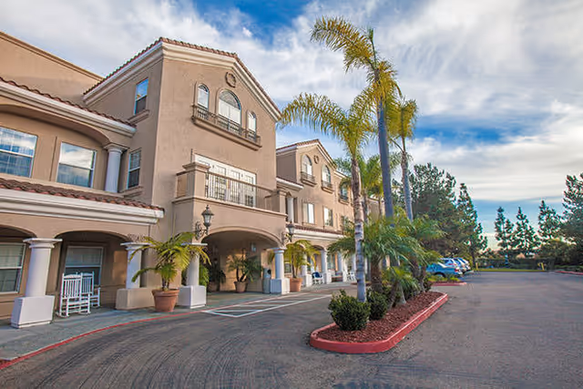 Front entrance of a three-story stucco building with palm trees, potted plants, rocking chairs, and a curved driveway.