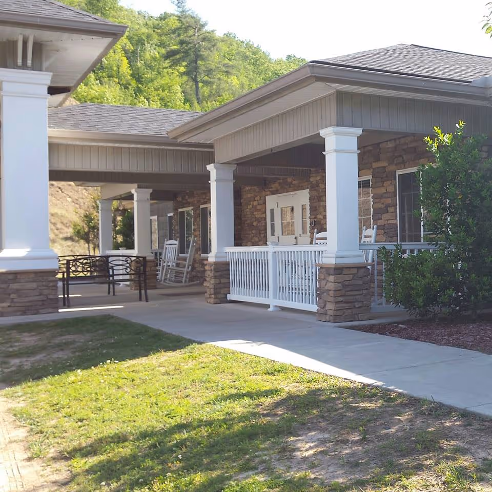 Covered porch area of a building with stone and white pillars, white railings, rocking chairs, and a bench, surrounded by greenery and trees in the background.