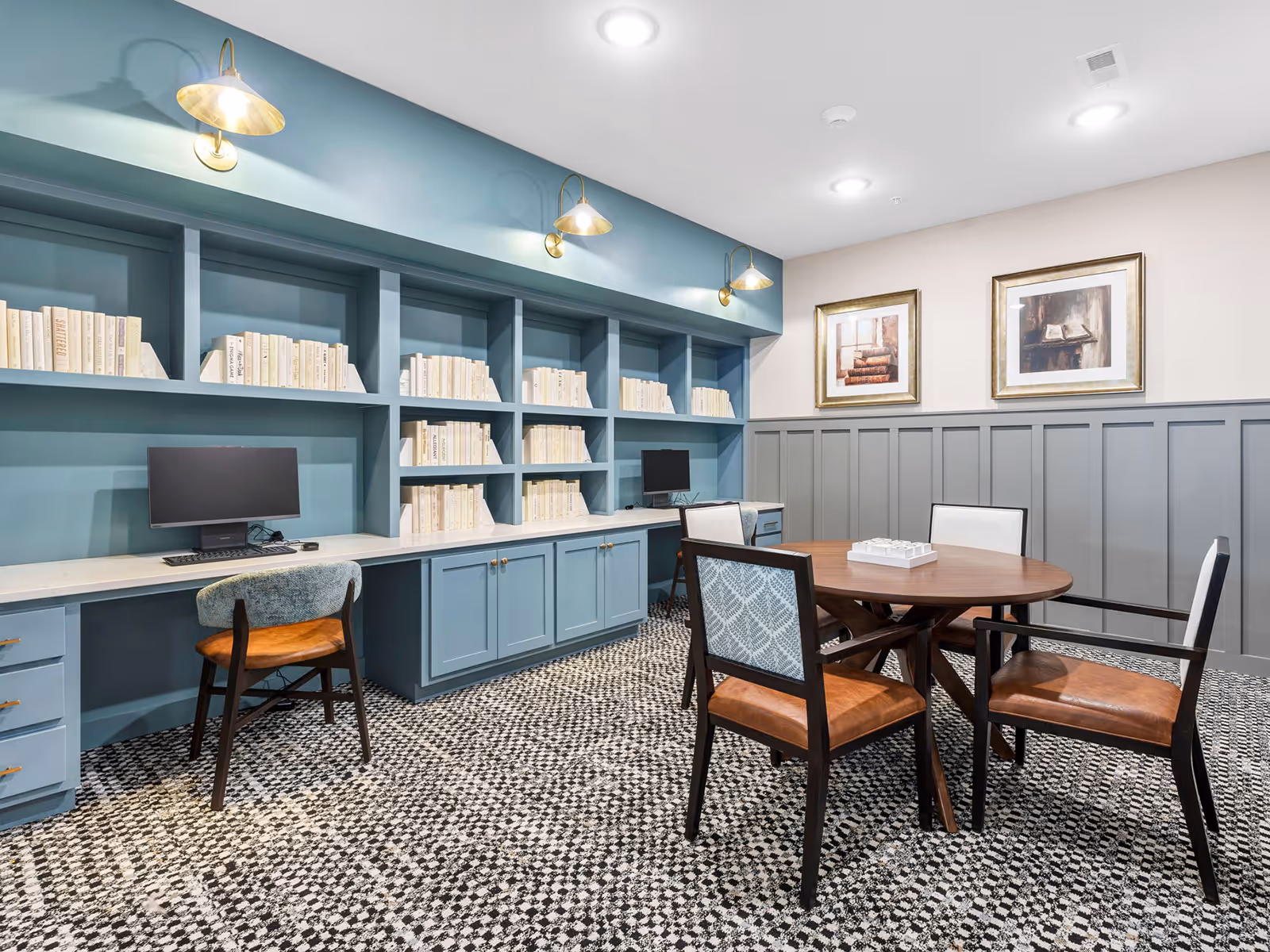 A cozy senior living facility room featuring a round wooden table with four chairs, two computer desks with monitors, built-in blue bookshelves filled with books, and two framed paintings on the wall. The room has patterned carpet flooring and soft lighting from wall-mounted lamps and ceiling lights.