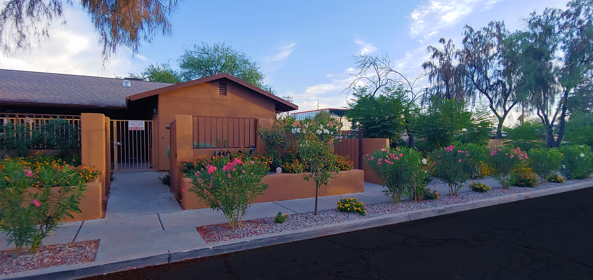 Exterior view of a single-story building with a brown facade and a gated entrance. The front area features a sidewalk bordered by landscaped flower beds with various green plants and colorful flowers. Trees and a partly cloudy sky are visible in the background.