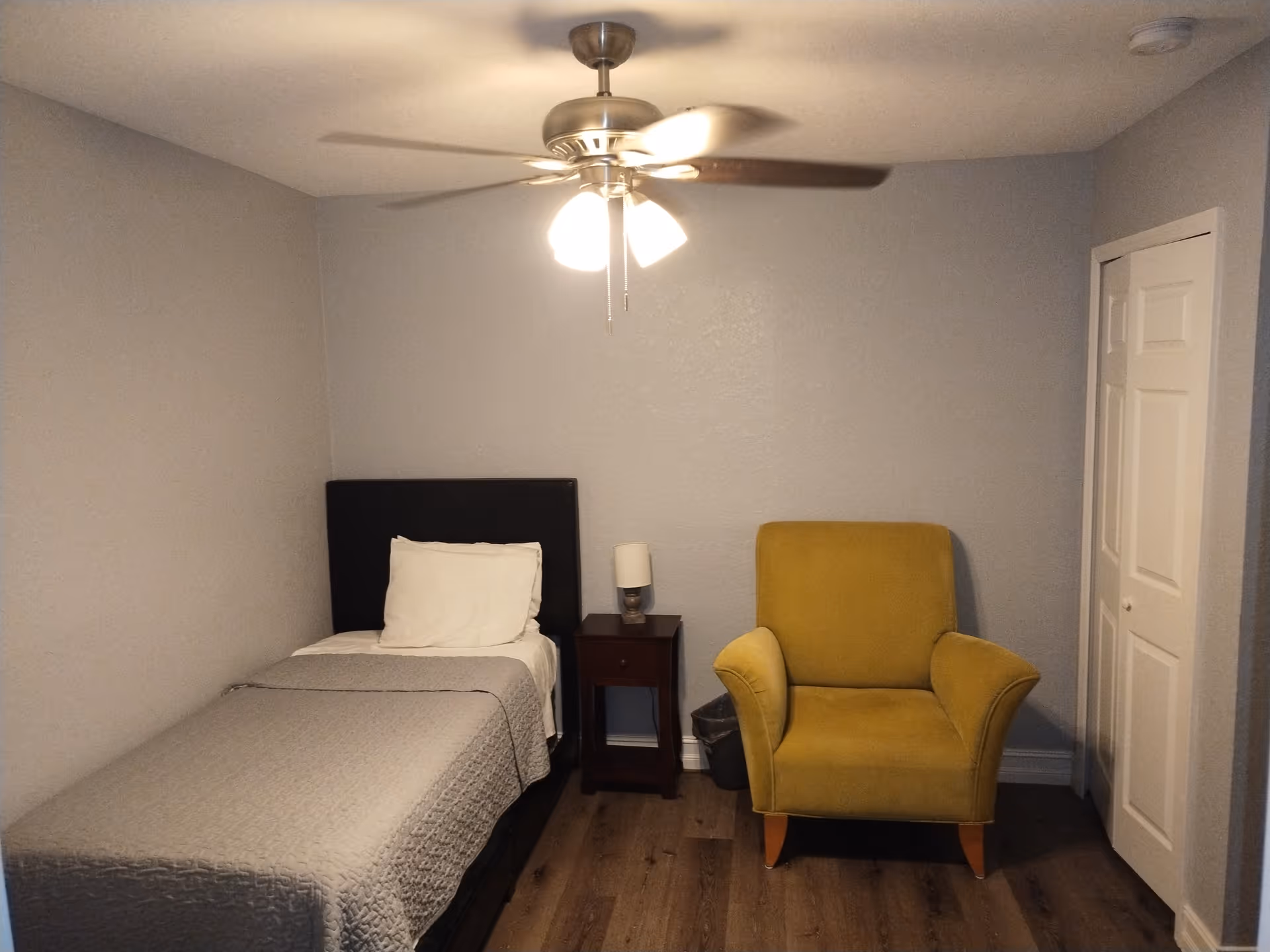 A small bedroom with a single bed covered with a gray quilt and a white pillow. Next to the bed is a dark wooden nightstand with a small lamp on it. To the right of the nightstand is a mustard yellow armchair. The room has light gray walls, a ceiling fan with lights, and a white closet door on the right side.