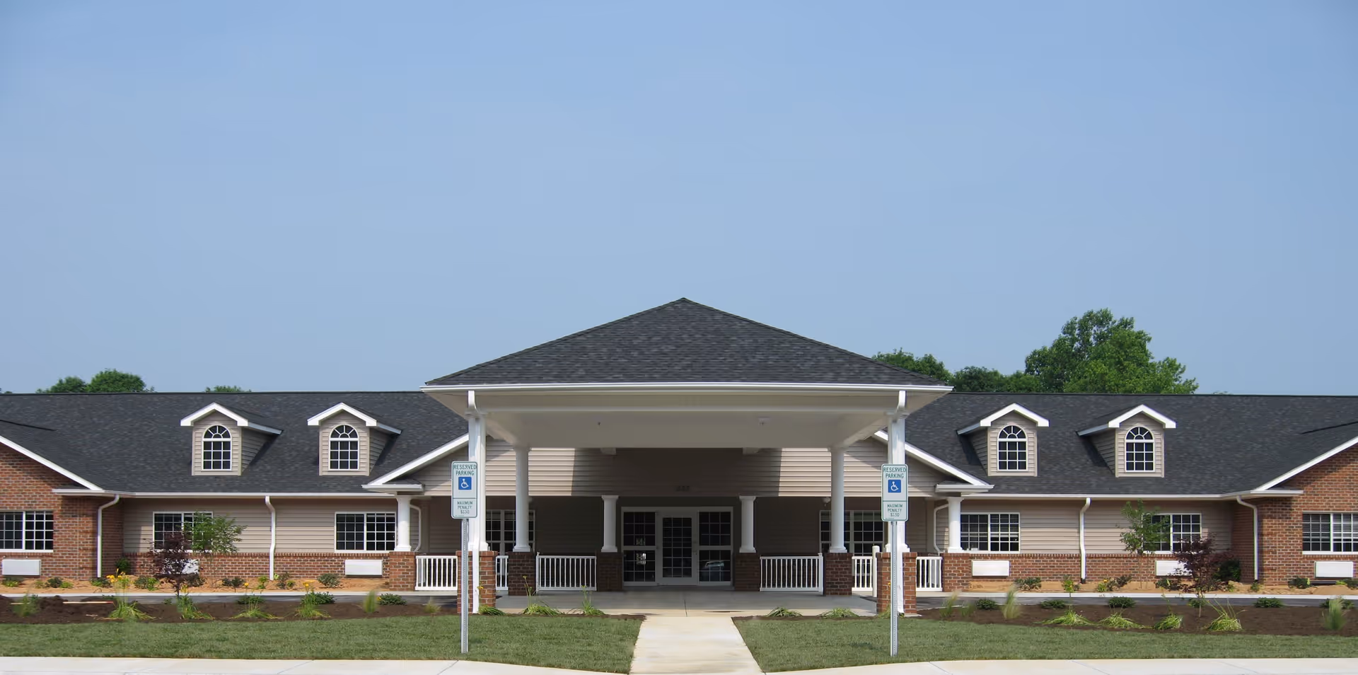 Front exterior view of Caswell House, a single-story building with a covered entrance supported by white columns, brick and siding facade, multiple windows, and a well-maintained lawn with two handicap parking signs near the entrance.