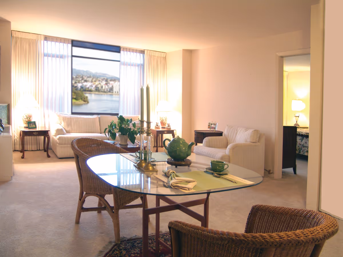 A cozy living room with a glass-top dining table set with green tea cups and a teapot, two wicker chairs, a white sofa and armchair, side tables with lamps, and a large window with sheer curtains showing a scenic view of water and buildings outside. An open doorway reveals a bedroom with a bed and lamp.