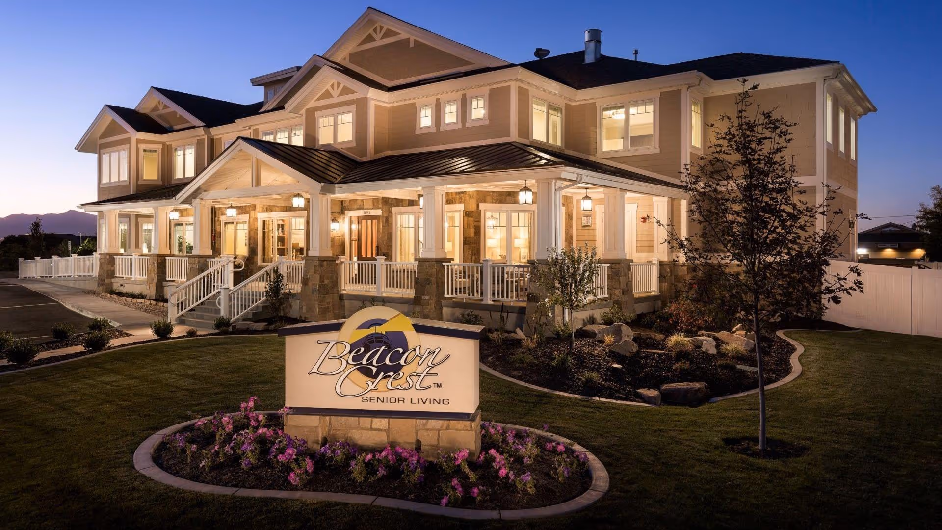 Exterior view of Beacon Crest Senior Living Draper building at dusk, showing a large two-story house-style structure with multiple windows lit from inside, a porch with white railings, landscaped garden with flowers and trees, and a sign in front displaying the facility's name.