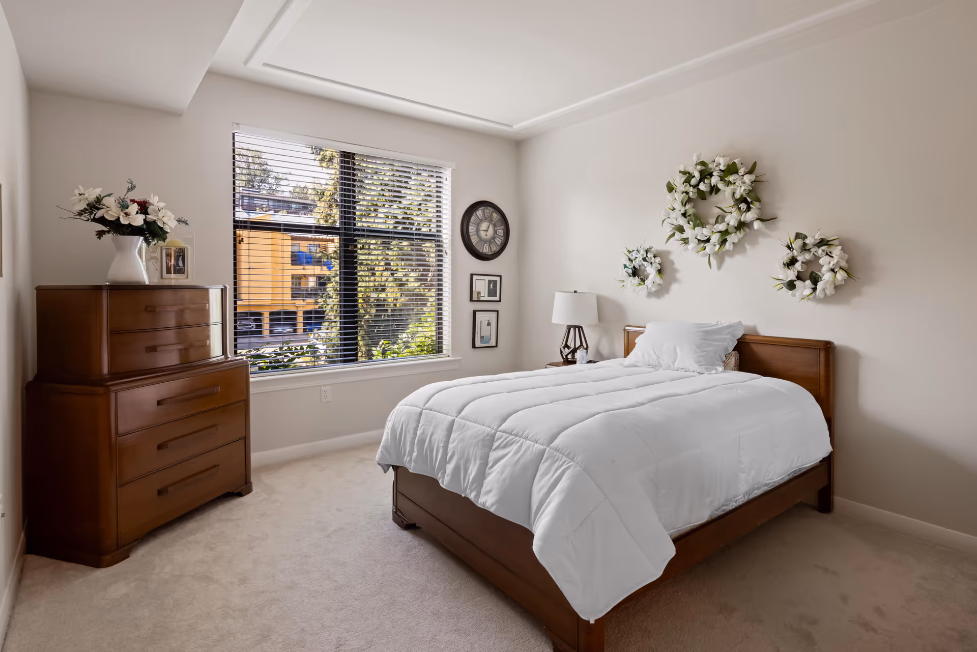Bright bedroom with a wooden bed and dresser, white bedding, floral wall wreaths, and a large window with blinds.