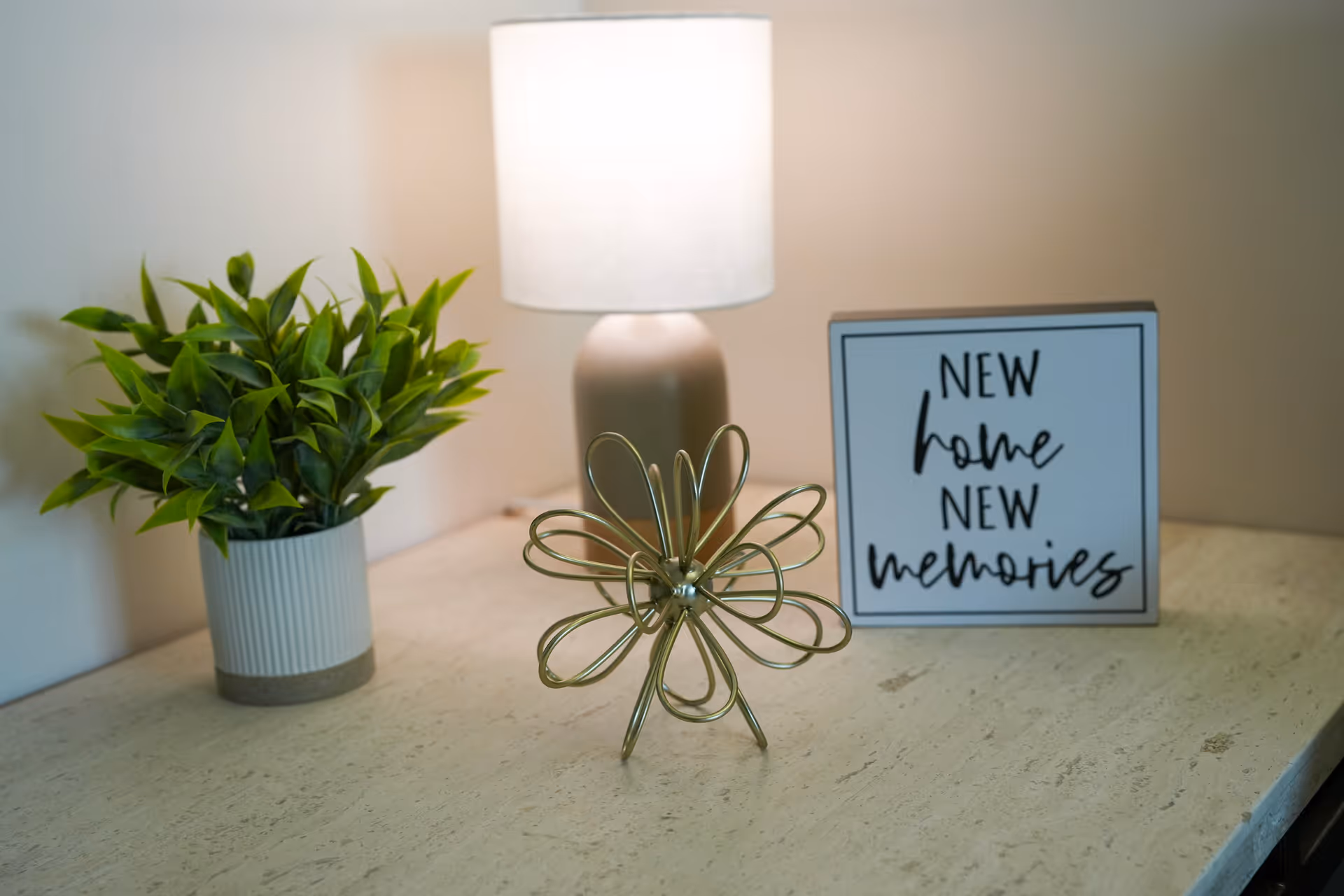 A tabletop with a small potted green plant, a beige lamp with a white lampshade, a decorative gold wire flower sculpture, and a framed sign that reads 'NEW home NEW memories'.