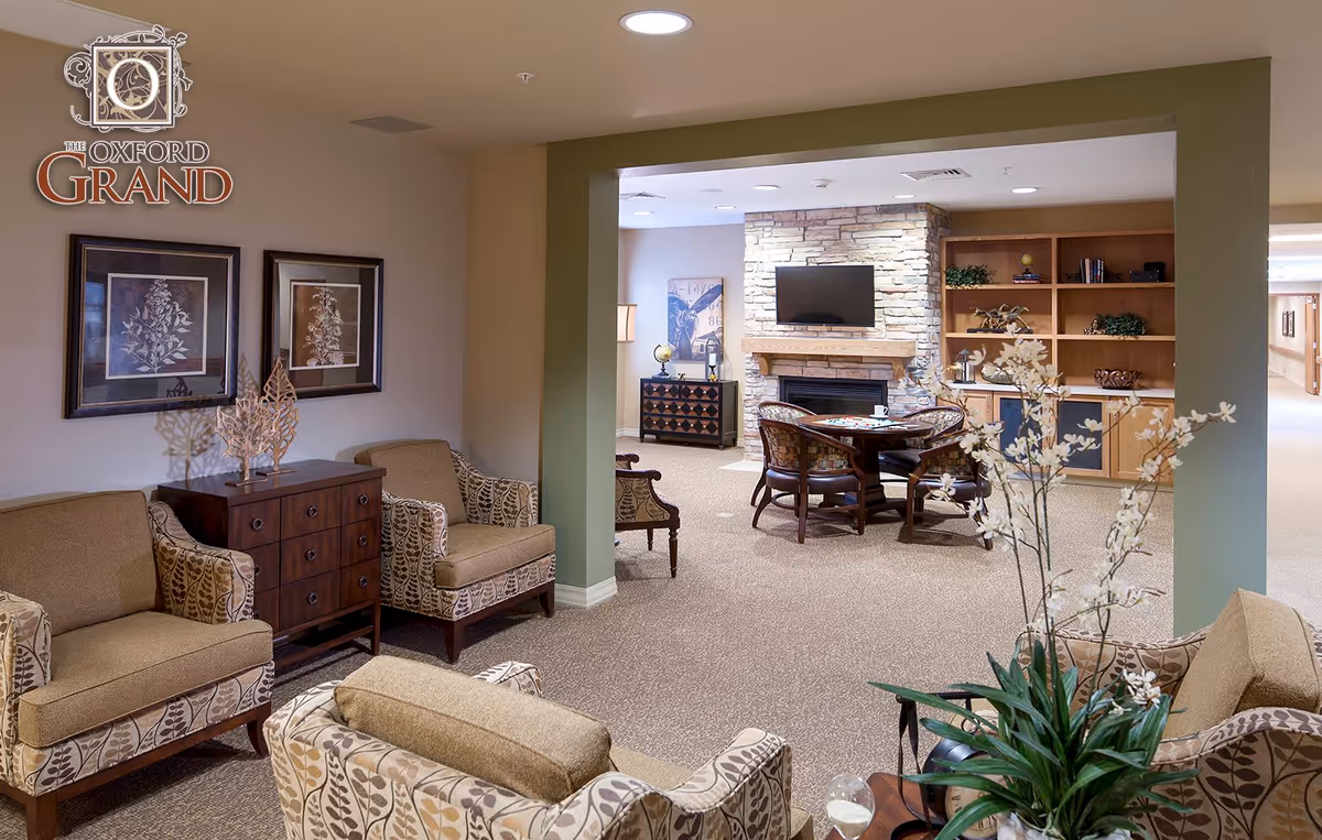 A cozy assisted living common area with patterned armchairs and a small sofa arranged around a wooden cabinet. In the background, there is a stone fireplace with a mounted flat-screen TV above it, a round table with chairs, and a wooden bookshelf with decorative items. The room is warmly lit with recessed ceiling lights.