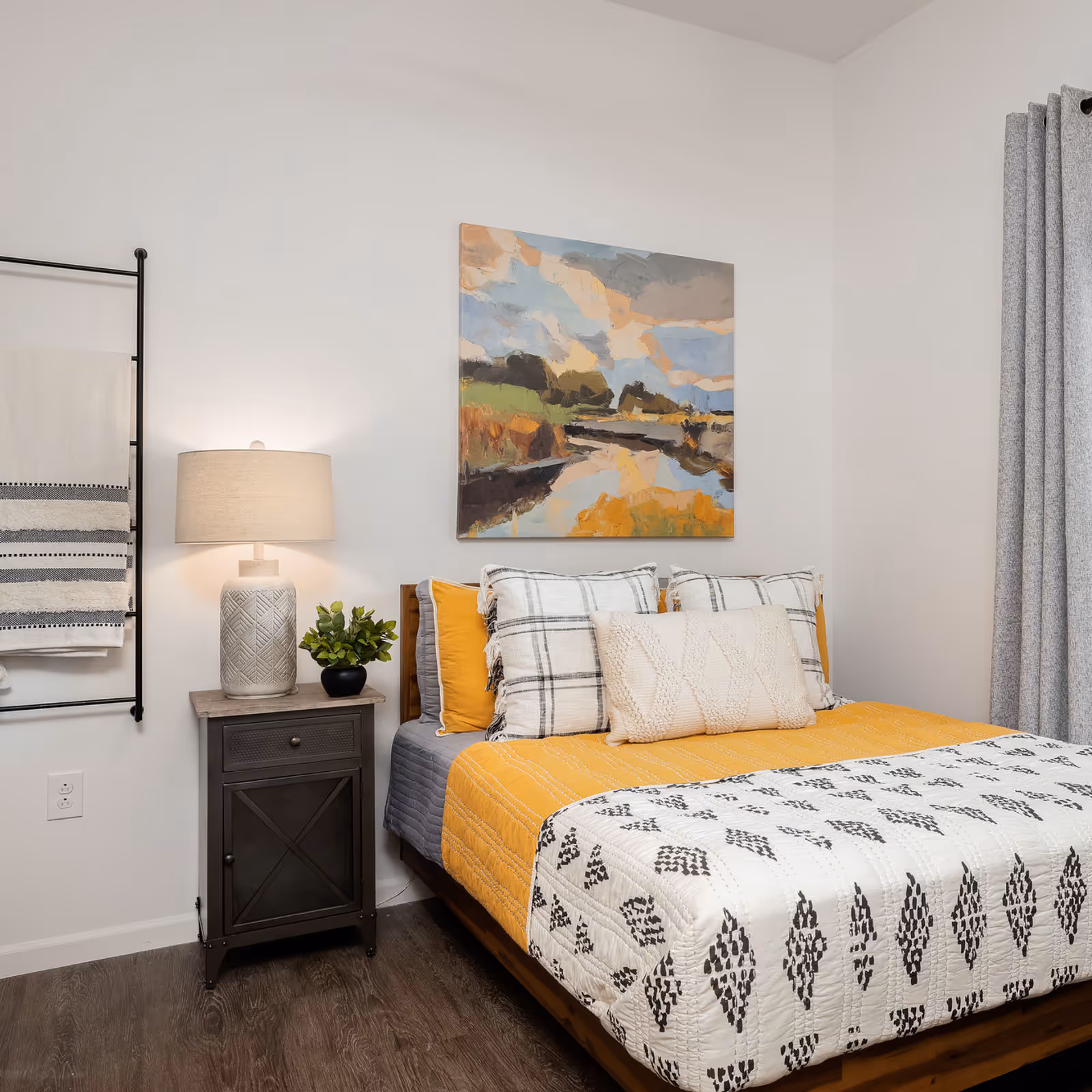 A cozy bedroom with a wooden bed frame, yellow and white bedding with black patterns, and multiple pillows. A nightstand with a decorative lamp and a small plant is beside the bed. Above the bed hangs a colorful abstract landscape painting. A towel rack with folded towels is mounted on the wall to the left, and gray curtains hang on the right side of the room.
