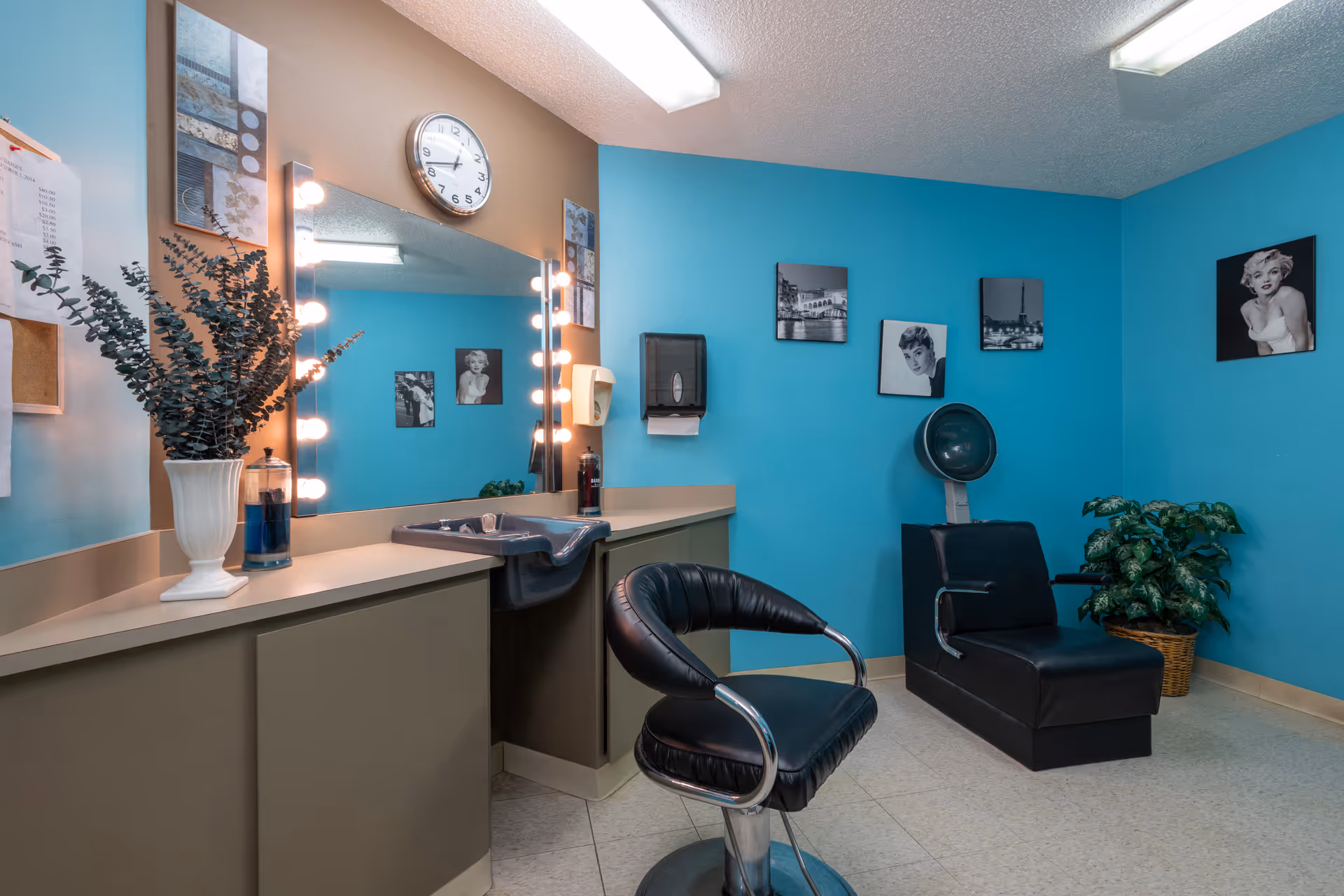 Interior of a salon room with blue walls, featuring a large mirror with lights around it, a clock above the mirror, a black salon chair in front of a counter with a sink, and a black hair drying chair. The walls are decorated with black and white photos and there is a potted plant in the corner.