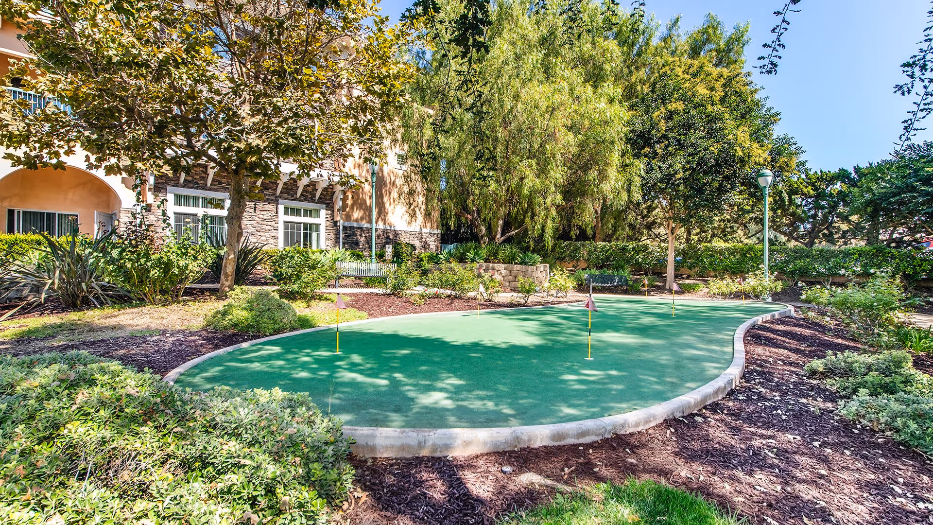 A putting green surrounded by landscaped garden beds with shrubs and trees, located next to a residential building with windows and stone accents under a clear blue sky.