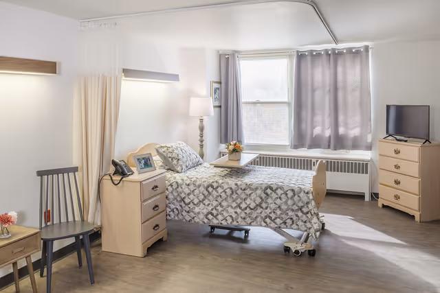 A bright furnished senior bedroom with a single adjustable bed, bedside tables, chair, lamp, window with curtains and a dresser with a TV.