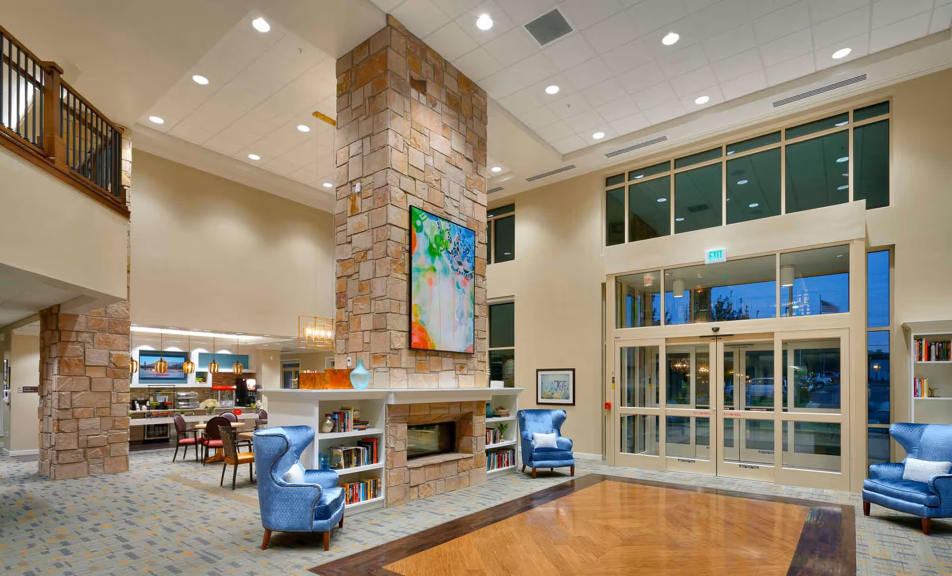 Spacious and well-lit senior living facility common area with a tall stone fireplace in the center, two blue armchairs with white pillows, bookshelves built into the fireplace base, large windows and glass doors letting in natural light, and a dining area with tables and chairs visible in the background.