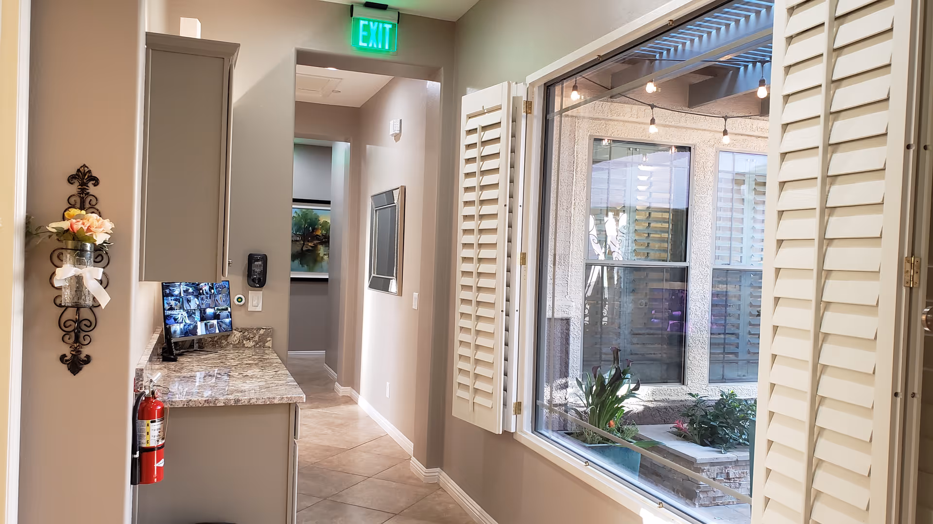 Interior view of a hallway in a senior living facility with beige walls and tiled floor. On the left side, there is a granite countertop with cabinets above and a fire extinguisher mounted below. A small monitor displaying security camera feeds is on the countertop. A decorative wall piece with flowers is mounted on the wall. On the right side, there is a large window with white plantation shutters looking out to an outdoor patio area with plants and string lights. An illuminated green exit sign is visible above the hallway entrance.