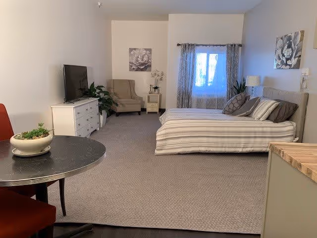 A spacious bedroom in an assisted living facility featuring a bed with striped bedding, a nightstand with a lamp, a cushioned armchair, a dresser with a flat-screen TV, a round table with a plant, and a window with patterned curtains.