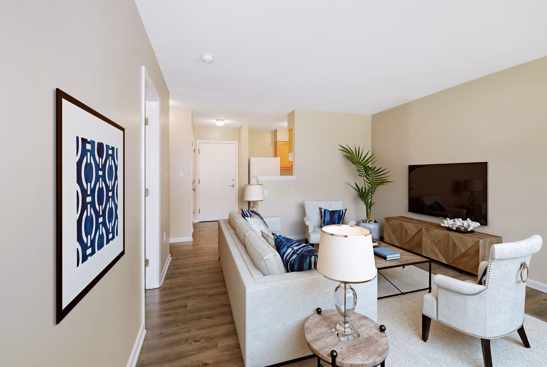 A modern living room in a senior apartment featuring a beige sofa with blue patterned pillows, two light-colored armchairs, a wooden coffee table, and a wooden TV stand with a flat-screen television mounted on the wall. There is a framed abstract blue and white artwork on the left wall, a small round side table with a lamp, and a potted plant near the TV stand. The room has light beige walls and wood flooring, with a view into the kitchen area and the apartment entrance door in the background.