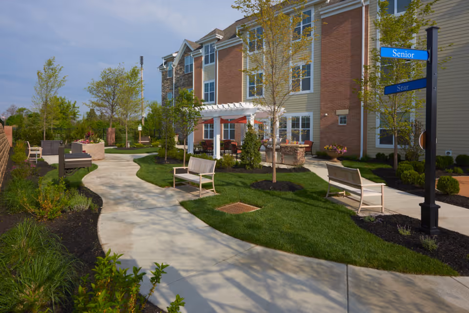 Outdoor garden area at Dublin Retirement Village featuring a winding concrete pathway, green grass, several benches, young trees, and a building with brick and siding in the background. There is a pergola with seating underneath and a signpost with blue signs reading 'Senior' and 'Star'.
