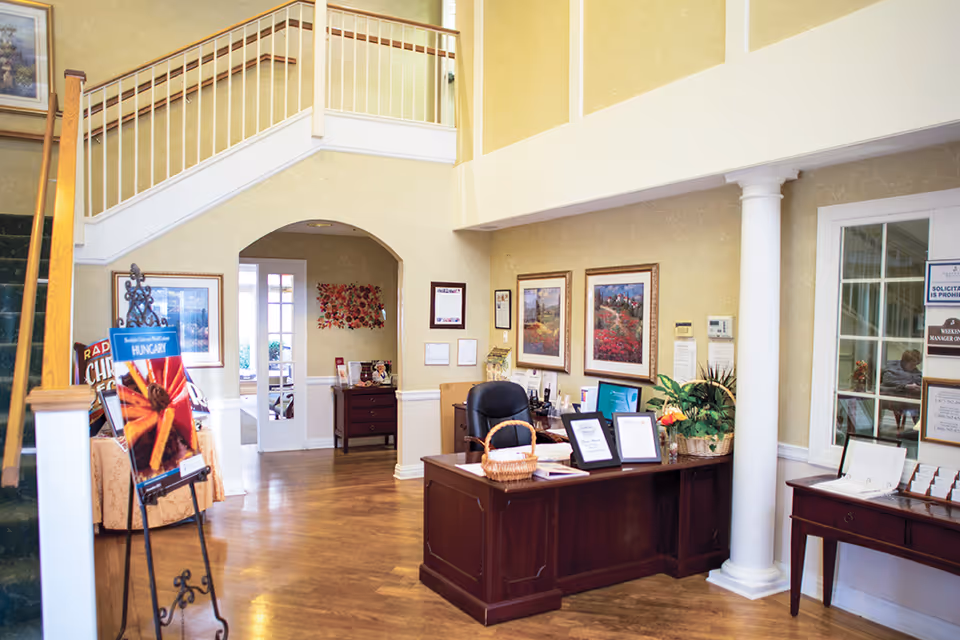 Reception area inside a senior living facility with a wooden desk, office chair, framed pictures on the wall, a basket on the desk, and a staircase with green carpet leading upstairs. There is a doorway leading to another room and a small table with brochures and decorations.