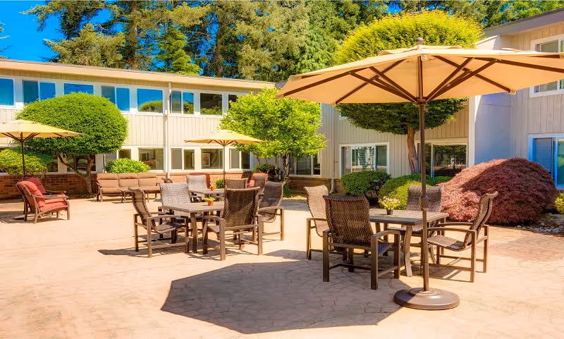 Outdoor patio area at Sagebrook Senior Living at Bellevue with several tables and chairs under large beige umbrellas, surrounded by well-maintained bushes and trees, with a two-story building in the background under a clear blue sky.