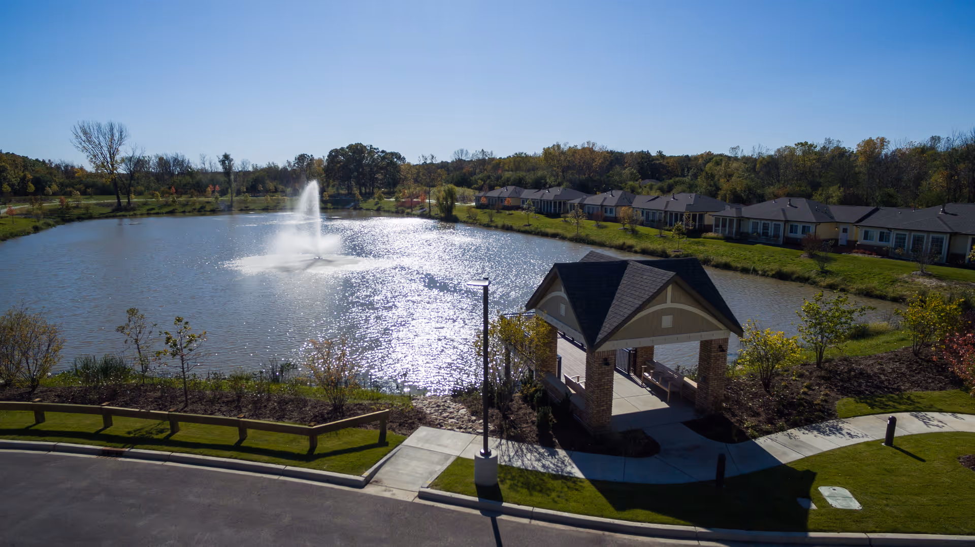 A scenic outdoor view of a senior living facility featuring a large pond with a water fountain in the center, surrounded by green lawns and trees. A covered seating area with benches is visible near the pond, and a row of single-story residential buildings lines the far side of the pond under a clear blue sky.