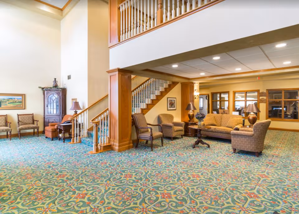 A spacious and well-lit senior living facility common area with patterned carpet, wooden staircase with white spindles, and comfortable seating including armchairs and a sofa around a wooden coffee table. The walls are painted in warm tones and decorated with framed artwork and lamps.