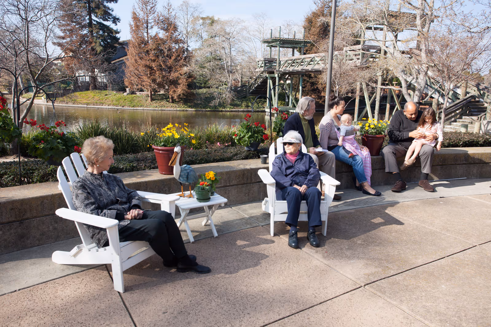 Seniors and family members sitting on outdoor chairs and a low wall beside a pond with flowers and a wooden walkway in the background.