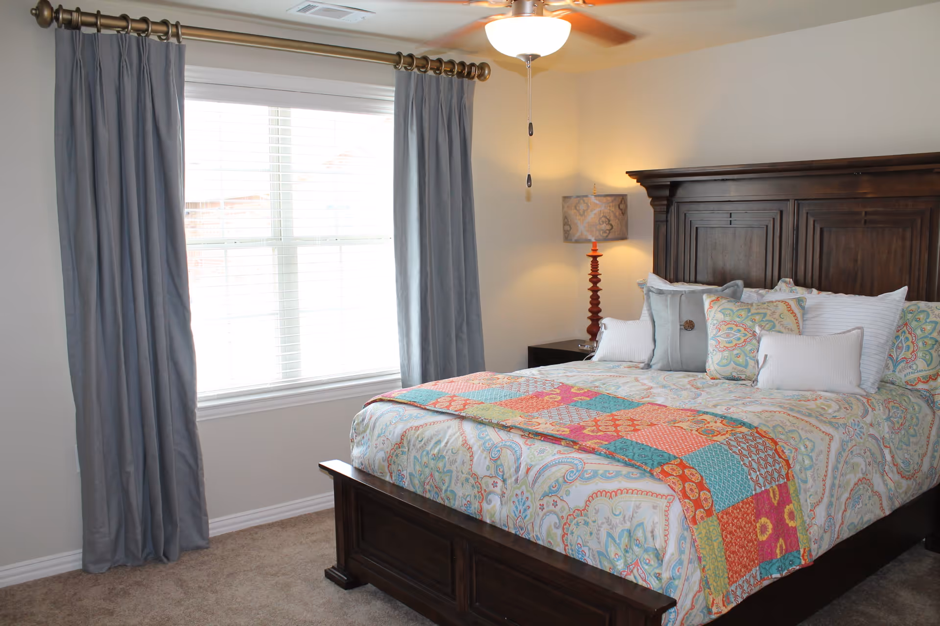 A bedroom with a large wooden bed frame and a colorful patterned bedspread. The bed is adorned with multiple decorative pillows. There is a window with closed blinds and gray curtains on the left side. A ceiling fan with a light is on the ceiling, and a bedside table with a lamp is visible in the corner.
