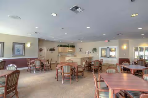 A spacious dining room with multiple wooden tables and chairs arranged neatly. The room has beige walls with framed artwork, soft lighting from ceiling fixtures and wall sconces, and a small kitchen or serving area in the background with cabinets and a countertop. Large windows allow natural light to enter the room.