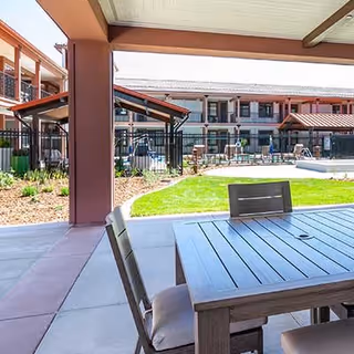 Covered outdoor patio area with a wooden table and cushioned chairs, overlooking a grassy lawn and a fenced swimming pool with a multi-story building in the background.