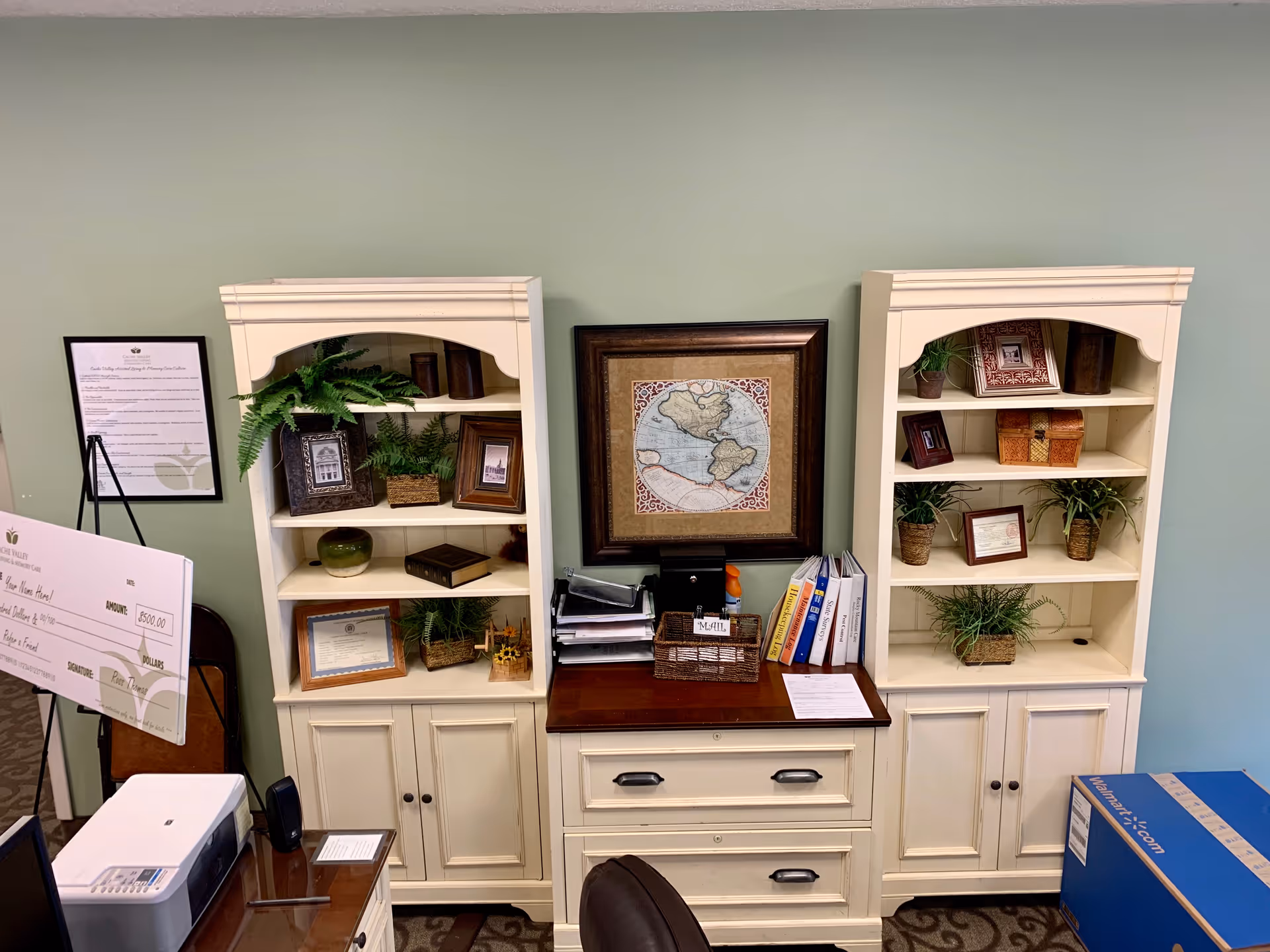 An office area with two white bookshelves filled with framed photos, plants, and decorative items. Between the bookshelves is a wooden desk with drawers, holding office supplies, folders, and a framed map on the wall above. A large check and a printer are visible on the left side, and a blue box is on the floor to the right.
