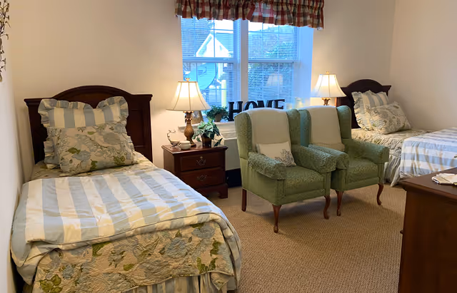 A cozy bedroom with two twin beds, each with striped and floral bedding. Between the beds is a wooden nightstand with a lamp, a small plant, and decorative items. In front of a window with a checkered valance are two green upholstered armchairs with white throws. The window has a decorative 'HOME' sign on the sill. The room has beige carpet and light-colored walls.