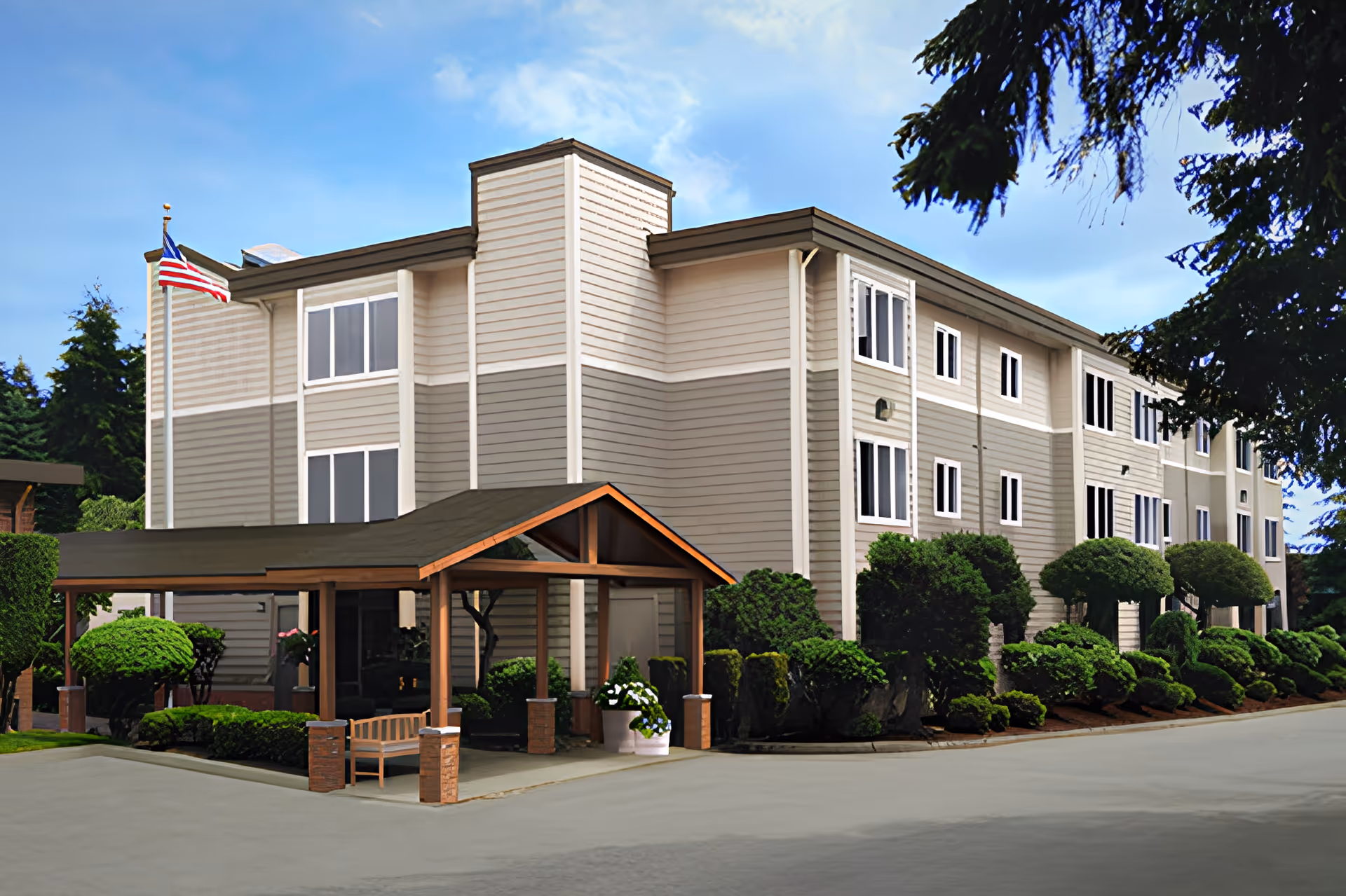 Exterior view of a three-story senior living facility building with beige and light gray siding, surrounded by well-maintained bushes and trees. There is a covered entrance with a wooden bench and an American flag flying on a flagpole near the building.
