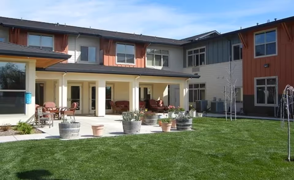 Outdoor view of a senior living facility courtyard with a well-maintained green lawn, several potted plants, patio seating with chairs and tables, and a two-story building with multiple windows in the background under a clear blue sky.