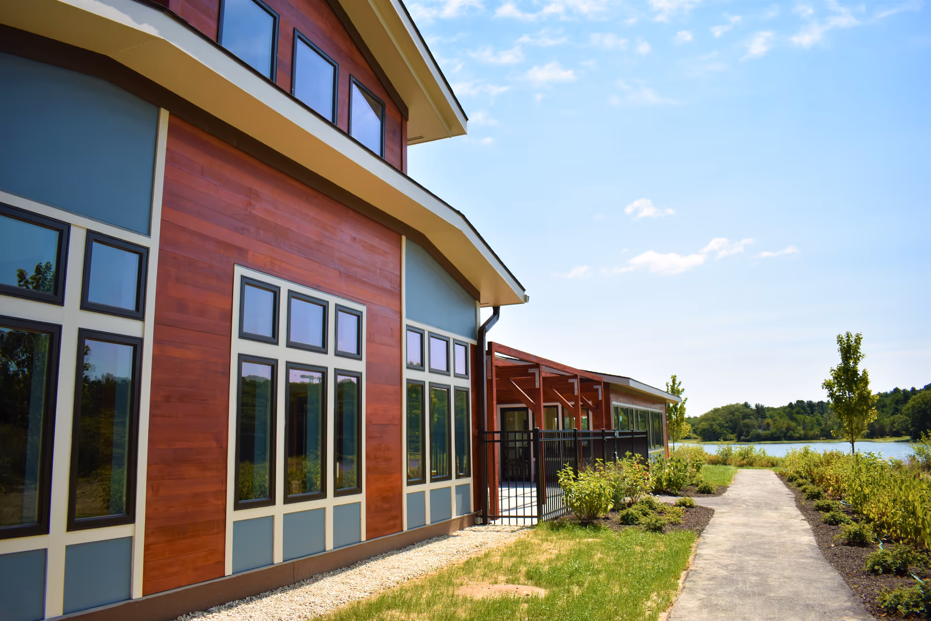 Exterior view of a modern building with large windows, wood paneling, and a pathway leading towards a lake surrounded by greenery under a blue sky with some clouds.