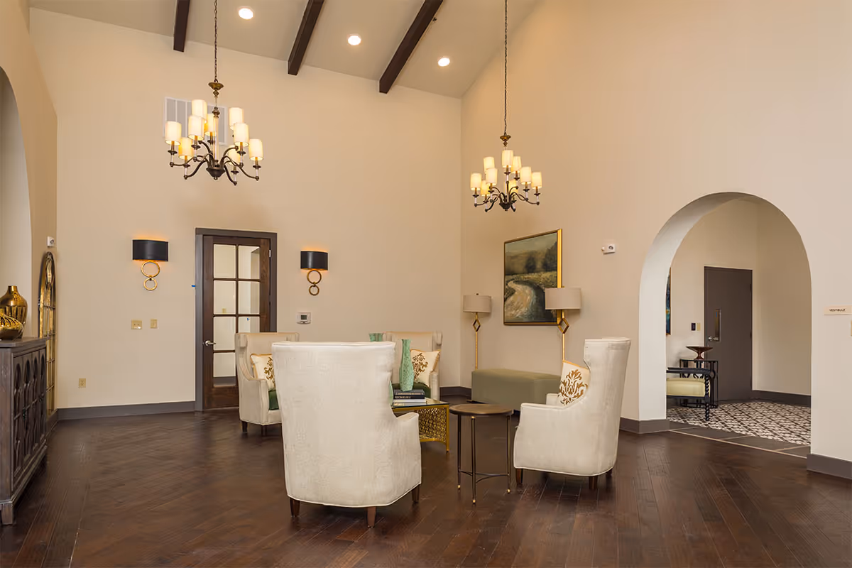 Well-lit lounge area with upholstered armchairs, side tables, chandeliers, and an arched doorway.