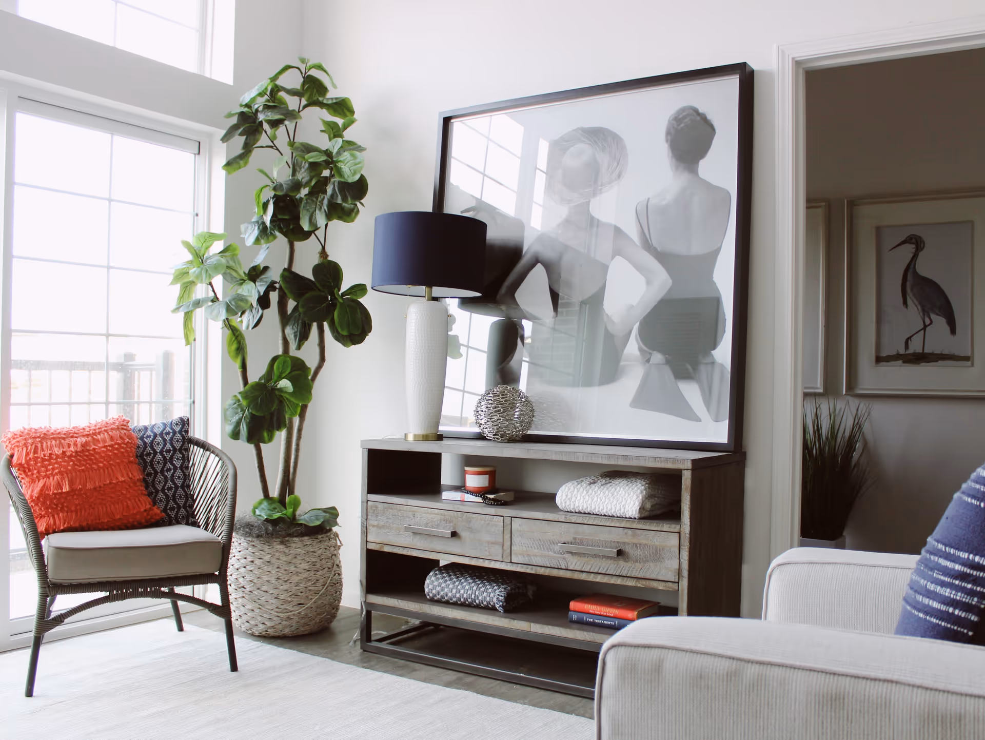 A bright living room area with a large window letting in natural light. There is a wicker chair with a coral and a navy blue pillow, a tall green potted plant in a woven basket, and a wooden console table with drawers. On the console table is a white lamp with a dark blue shade, a decorative silver sphere, folded blankets, and books. Above the console table is a large framed black and white photograph of two women seen from the back. In the background, there is a doorway leading to another room with framed artwork of a bird and a tall plant.