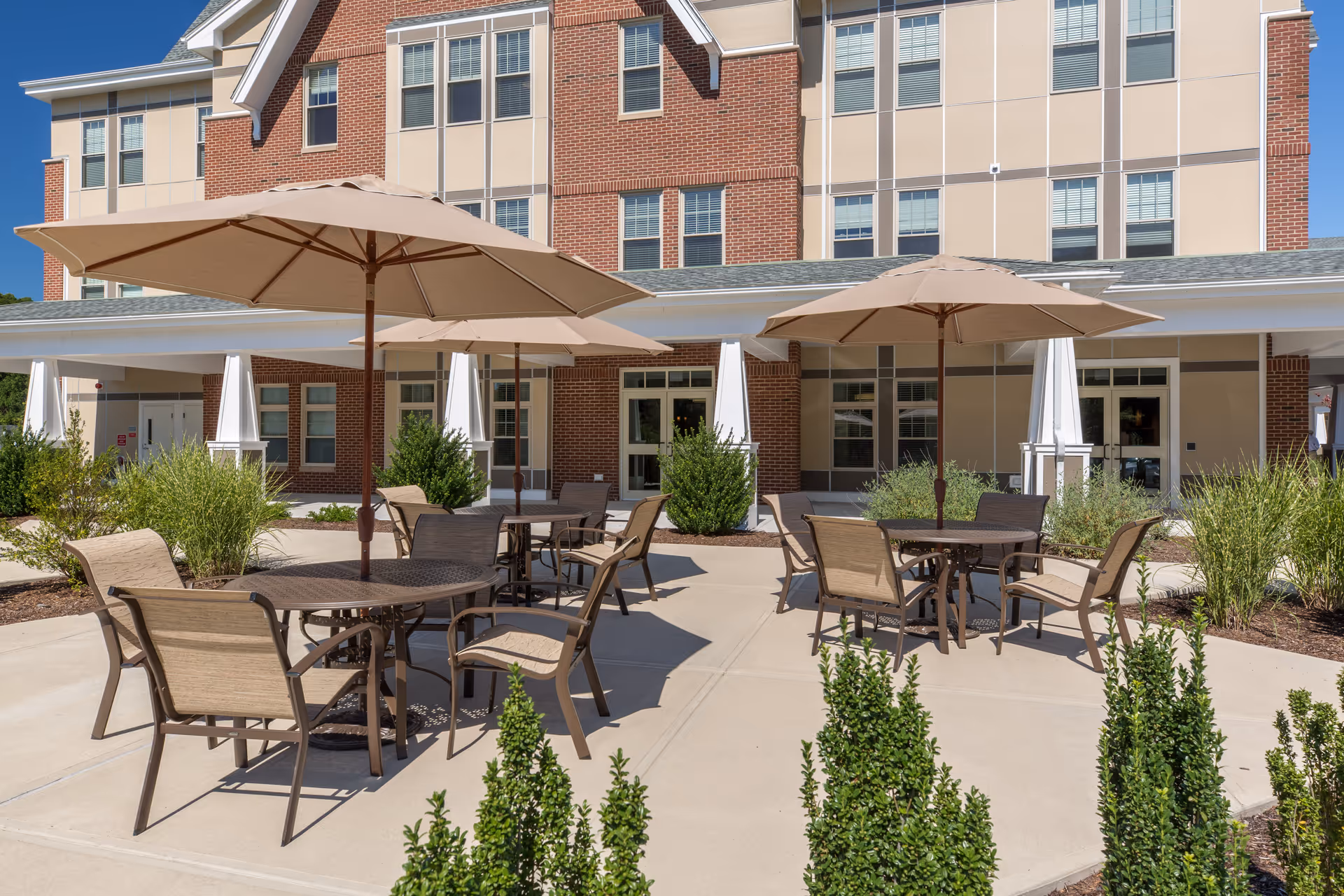 Outdoor patio area at The Residence at Valley Farm featuring several round tables with beige umbrellas and matching chairs, surrounded by landscaped greenery and a multi-story brick and beige building in the background under a clear blue sky.
