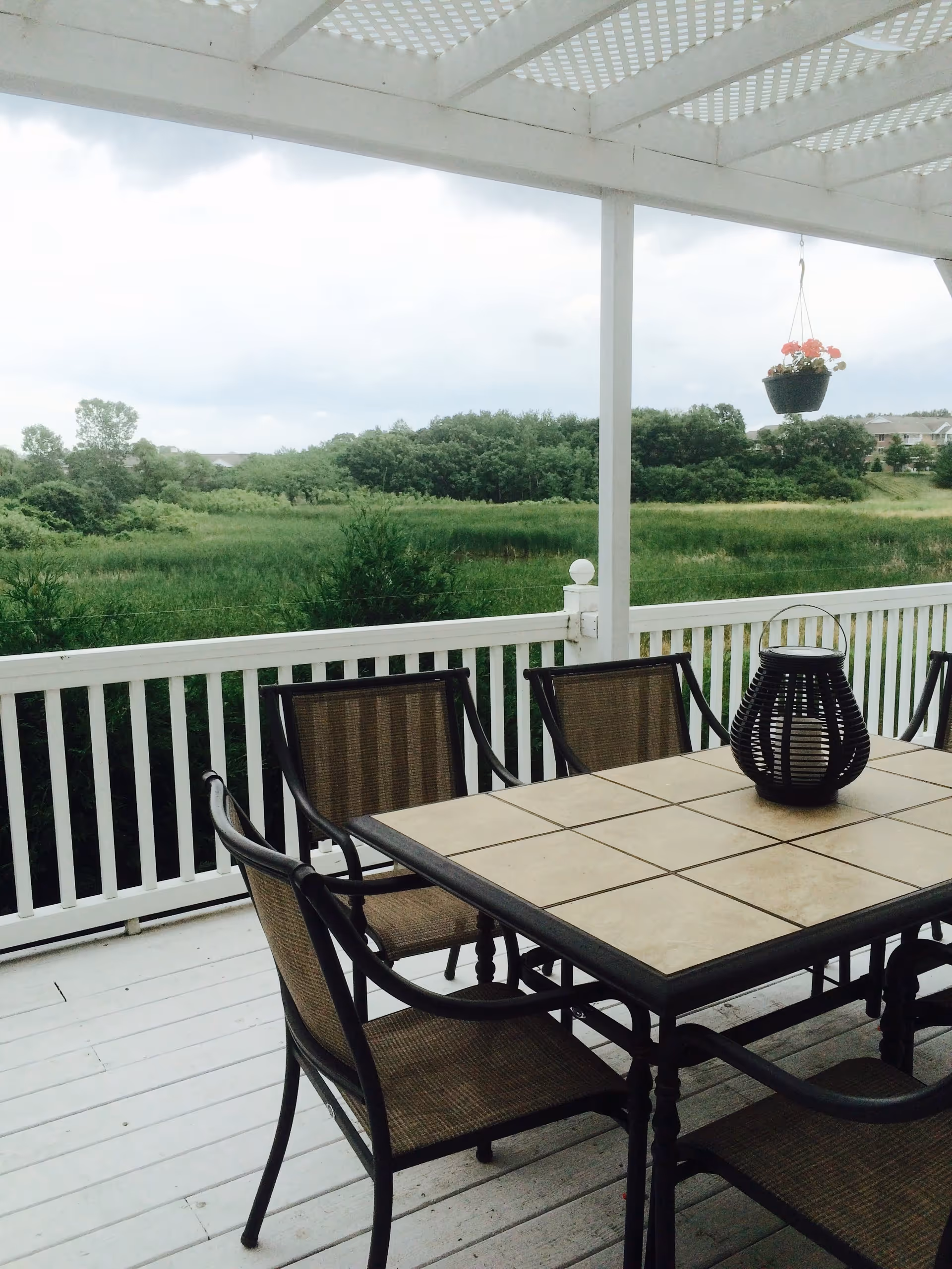 Outdoor patio area with a white wooden pergola overhead, a rectangular table with a tiled surface, surrounded by six chairs with mesh backs and seats. A black decorative lantern sits on the table, and a hanging flower pot with pink flowers is suspended from the pergola. In the background, there is a green field with trees and a cloudy sky.