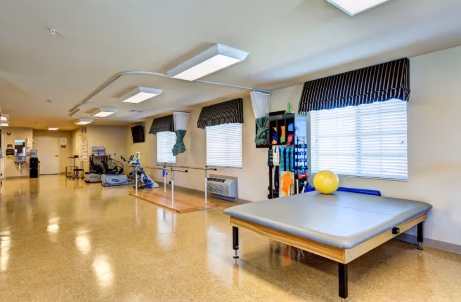 A rehabilitation therapy room with exercise equipment including stationary bikes, parallel bars for walking practice, a padded therapy table with a yellow exercise ball on it, and various therapy tools organized on a rack. The room has large windows with striped valances and bright overhead lighting.