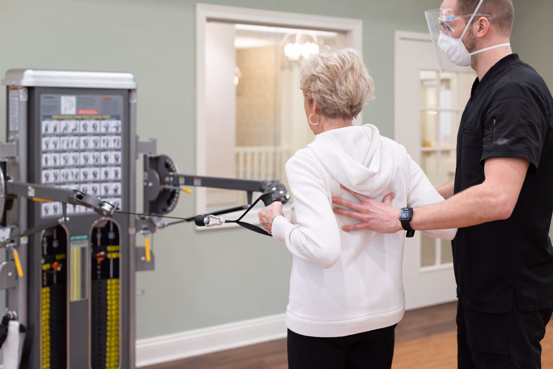 A senior woman in a white hoodie is exercising with resistance cables on a fitness machine, assisted by a male staff member wearing a black uniform, face mask, and face shield, in an indoor facility room with light green walls and wooden floors.