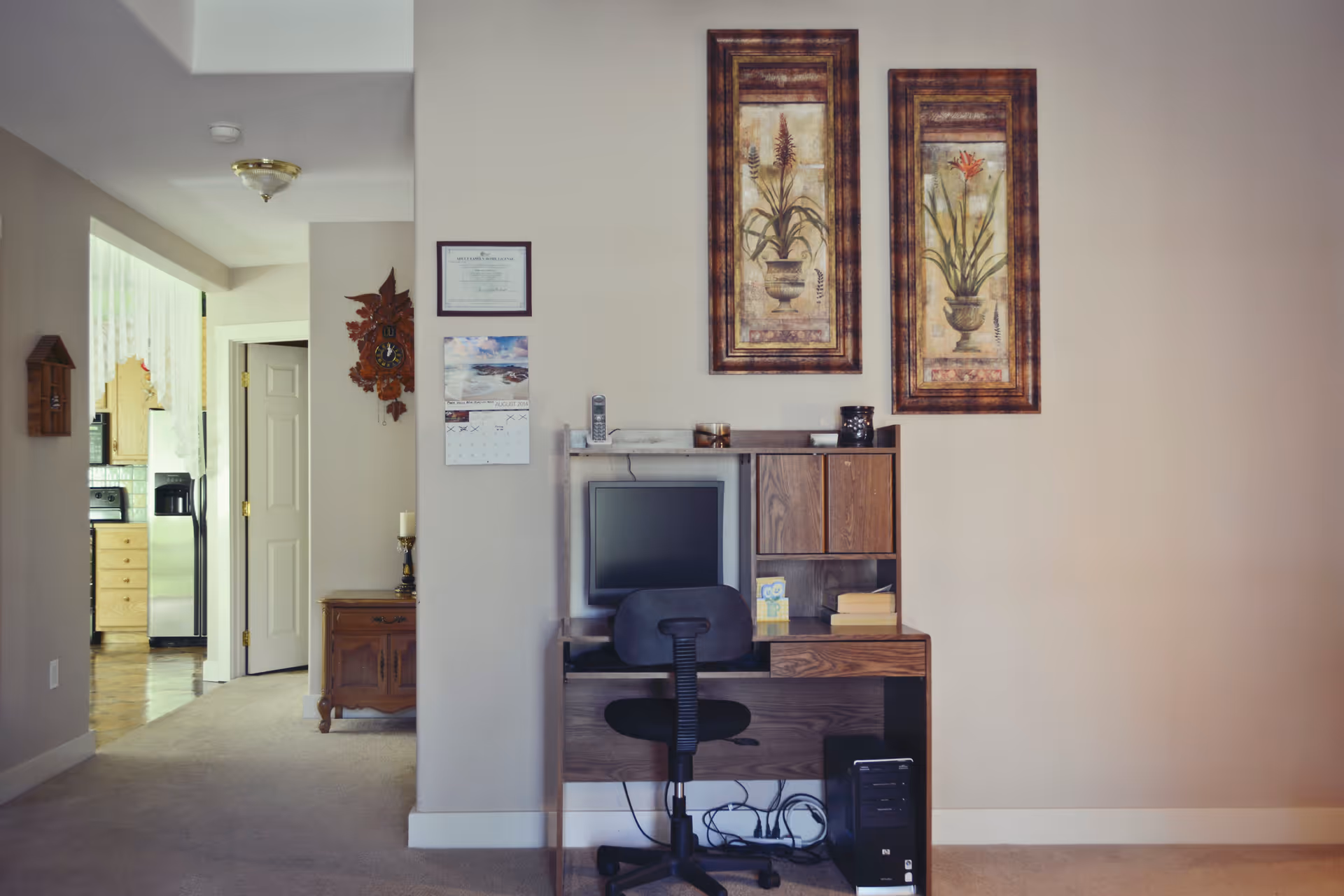 A small home office area with a wooden desk, a computer monitor, a black office chair, and a desktop computer tower underneath. Above the desk are two framed botanical prints. To the left, a hallway leads to a kitchen with wooden cabinets and stainless steel appliances. The walls are painted beige and there is a carpeted floor.