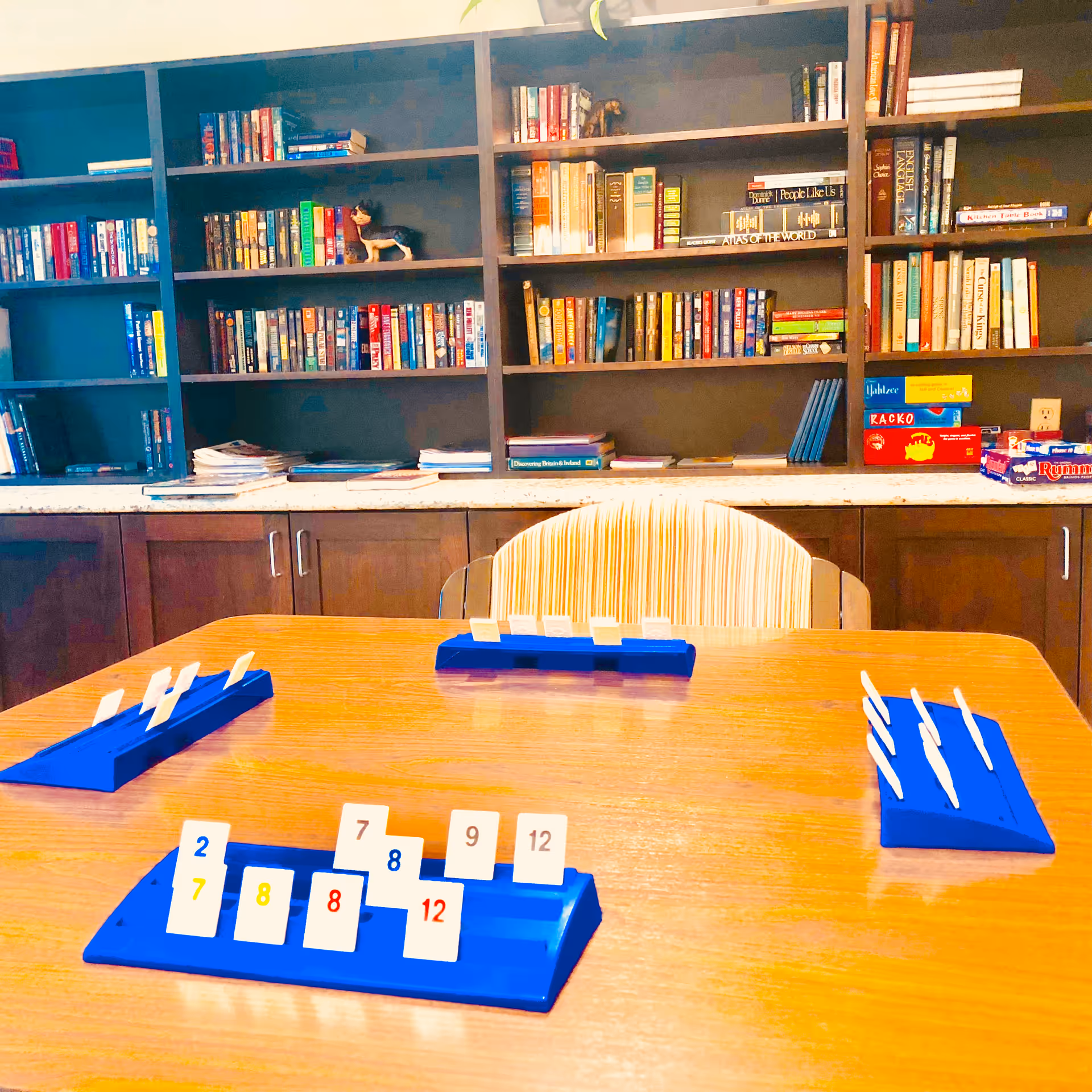 A game table with blue tile racks and numbered tiles in a senior living common room with bookshelves behind.