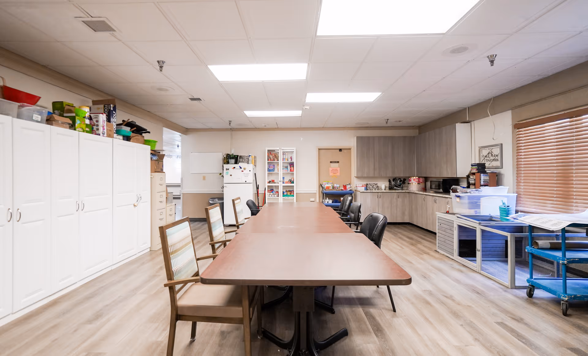 A large room with a long rectangular table surrounded by chairs, white storage cabinets on the left, a refrigerator and shelves with various items at the back, and a countertop with cabinets on the right. The room has wood flooring, a drop ceiling with fluorescent lights, and a window with wooden blinds on the right side.