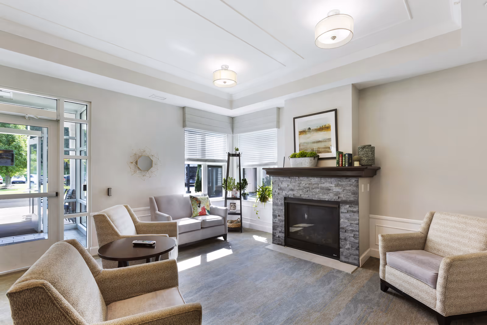 A bright and cozy living room with a stone fireplace, a framed painting above the mantel, and several potted plants. The room features a gray sofa with a decorative pillow, two beige armchairs, a round wooden coffee table with a remote control on it, large windows with blinds, and a glass door leading outside.