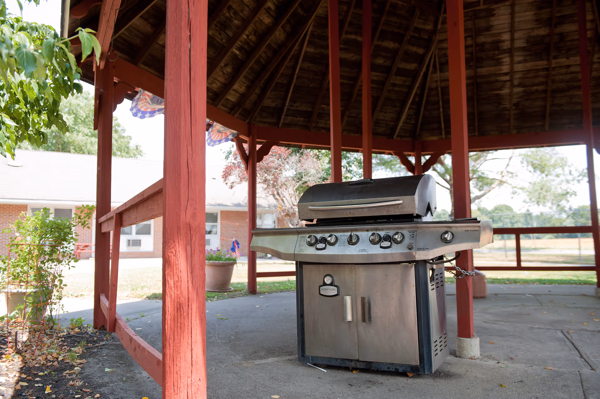 A stainless steel gas grill sits under a red wooden gazebo on a concrete pad with a brick building and trees in the background.