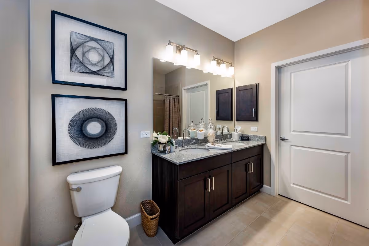 Modern bathroom featuring a toilet, dark-wood double-sink vanity with a large mirror, decorative framed art, and a closed white door.