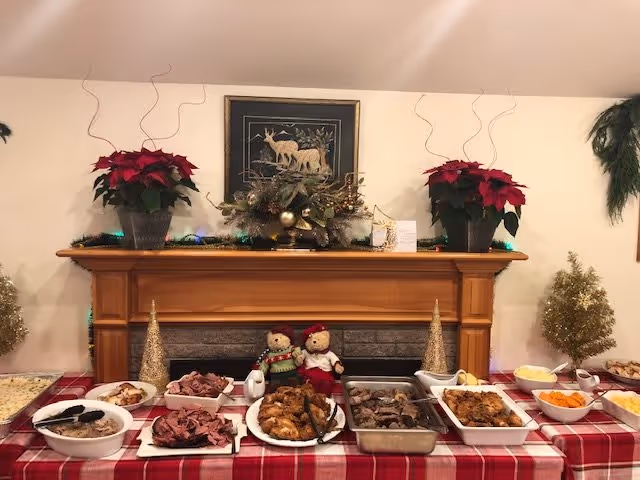 A buffet-style holiday meal setup on a table with a red and white checkered tablecloth in front of a wooden fireplace mantel decorated with poinsettia plants, holiday greenery, and a framed picture of deer. The table holds various dishes including sliced meats, fried chicken, vegetables, and other side dishes. Two teddy bears dressed in holiday attire are placed on the mantel.