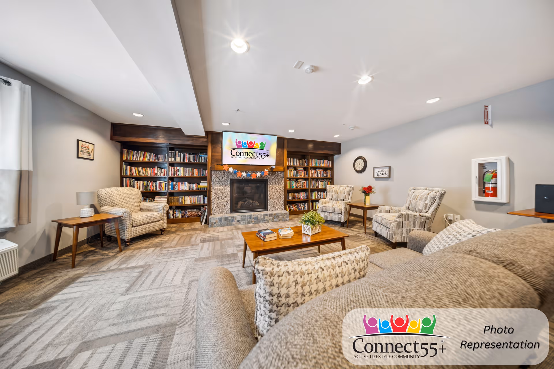 A cozy senior living community lounge with a fireplace surrounded by bookshelves filled with books. The room features comfortable armchairs and a sofa with patterned cushions, a wooden coffee table with books and a plant, and side tables with lamps and flowers. The walls are light gray, and there is a clock and a decorative sign on the wall. A television above the fireplace displays the Connect55+ logo.