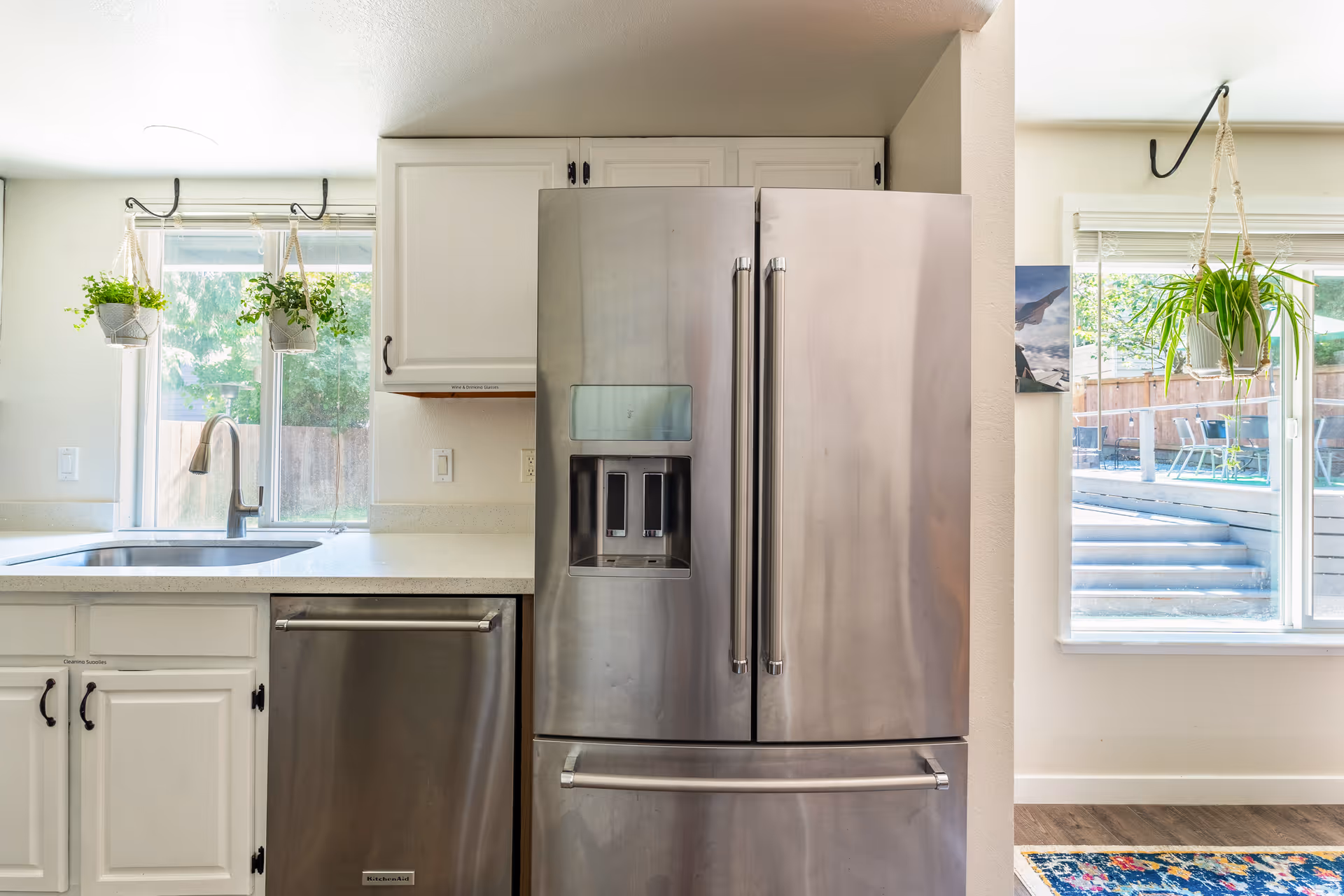 Modern kitchen area with a stainless steel refrigerator with French doors and a bottom freezer drawer, a dishwasher, white cabinetry, a sink under a window with two hanging potted plants, and a window showing an outdoor patio area with steps and chairs.